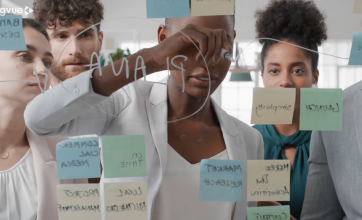 A business woman writes 'Plan A' on a clear board surrounded by post it notes as colleagues watch