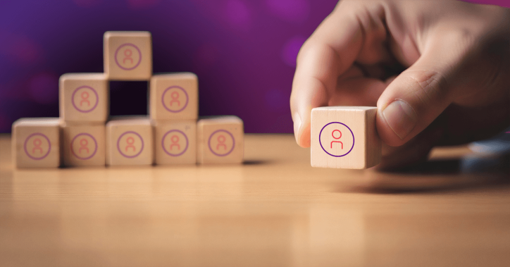 An image of some wooden blocks stacked on top of each other in the background, each with an icon representing a person. In the foreground, a hand grabs another block like this.