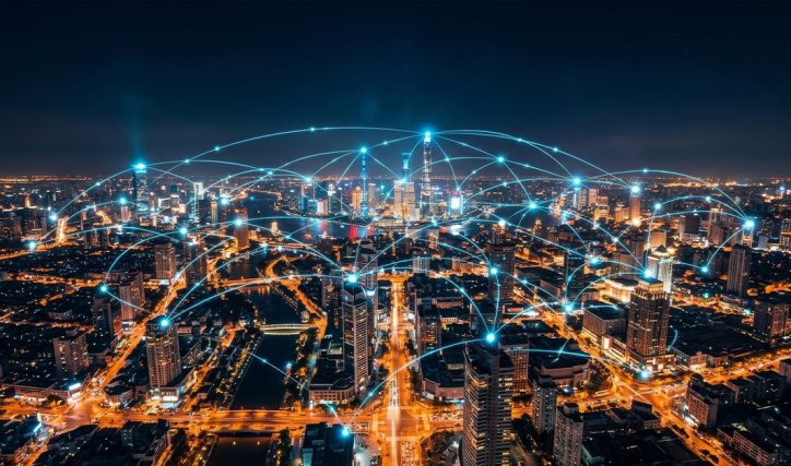 Aerial night view of a modern city skyline illuminated with lights, overlaid with glowing blue network connections linking buildings and districts to represent digital connectivity, data exchange, and smart city infrastructure.